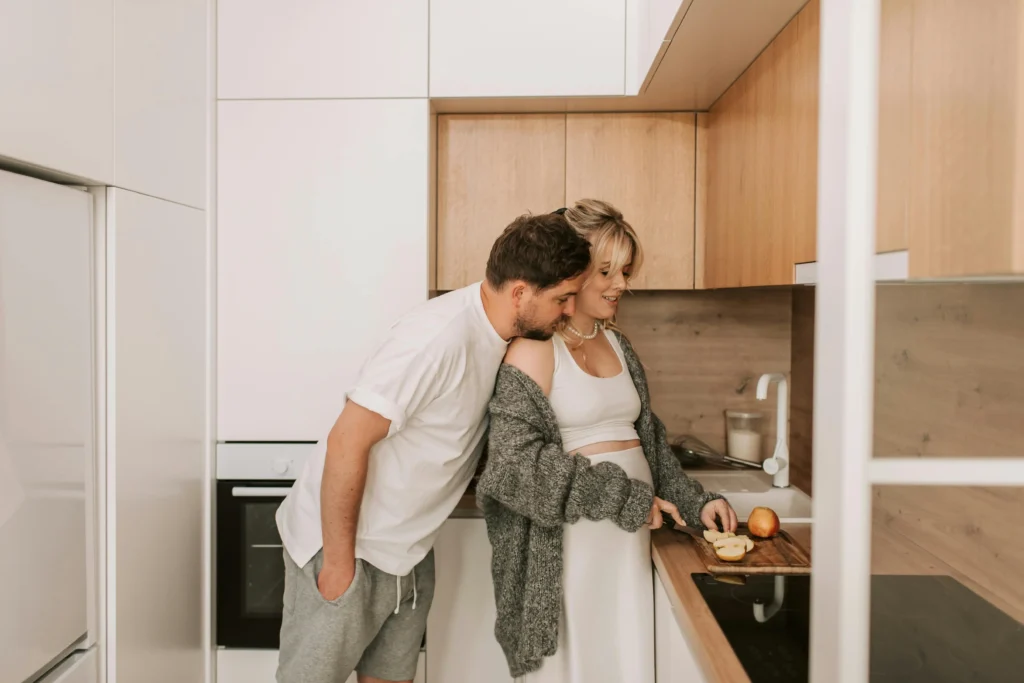 Kobieta w ciąży kroi jabłko w kuchni, partner czule obejmuje ją i całuje w ramię