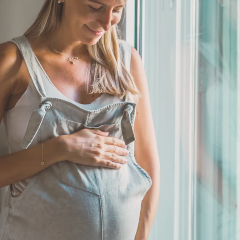 Uśmiechnięta kobieta w ciąży dotyka brzucha, stojąc przy oknie w naturalnym świetle