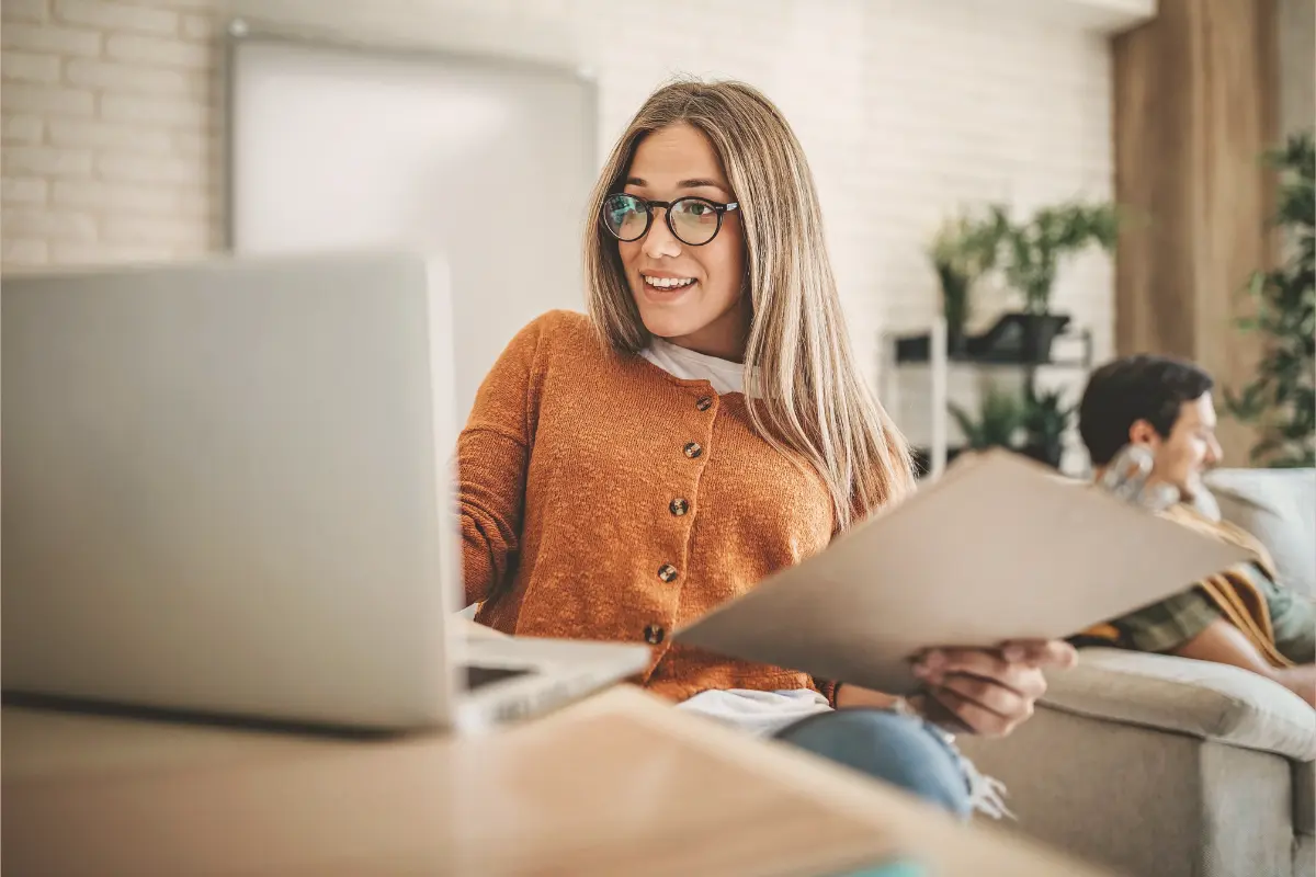 Uśmiechnięta kobieta uczy się przy laptopie w salonie, w tle partner na kanapie