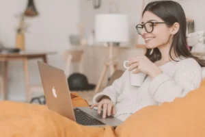Uśmiechnięta kobieta w okularach uczy się przy laptopie z kubkiem kawy, wygodnie otulona kocem