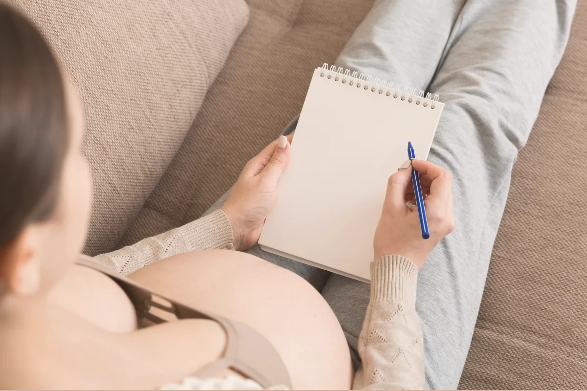 Kobieta w ciąży siedzi na kanapie i notuje w notesie swoje preferencje do planu porodu