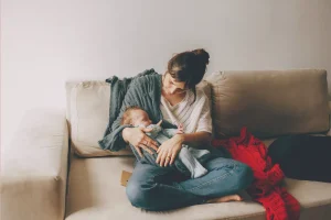 Mama siedzi na kanapie w domu i trzyma śpiącego noworodka, czekając na wizytę położnej